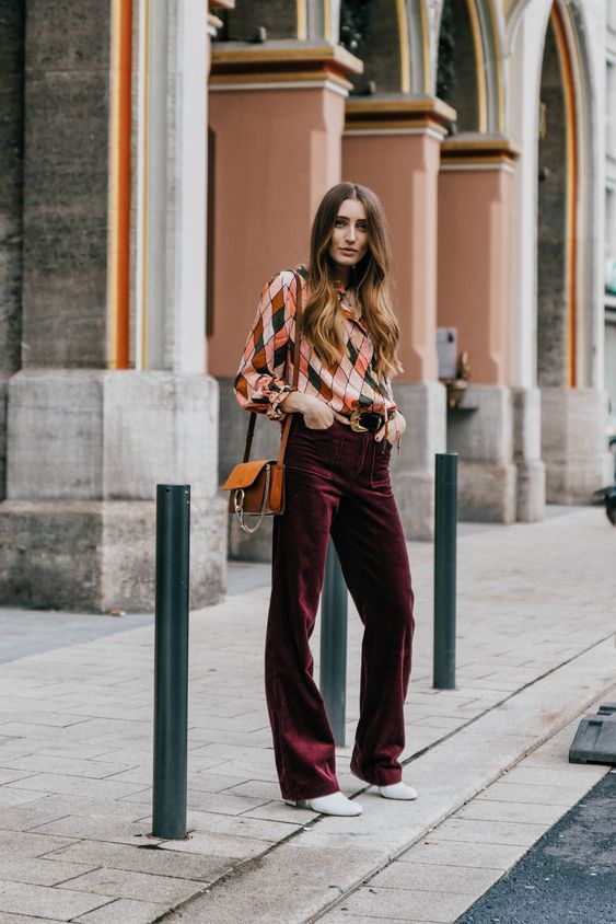 Bold and cool wedding guest look with a colorful printed top, burgundy velvet pants, white shoes, and an amber bag