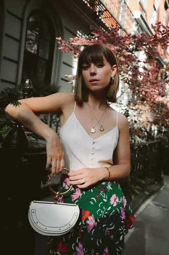 Chic outfit with a button-up spaghetti strap top, bright floral skirt, white bag, and layered necklaces