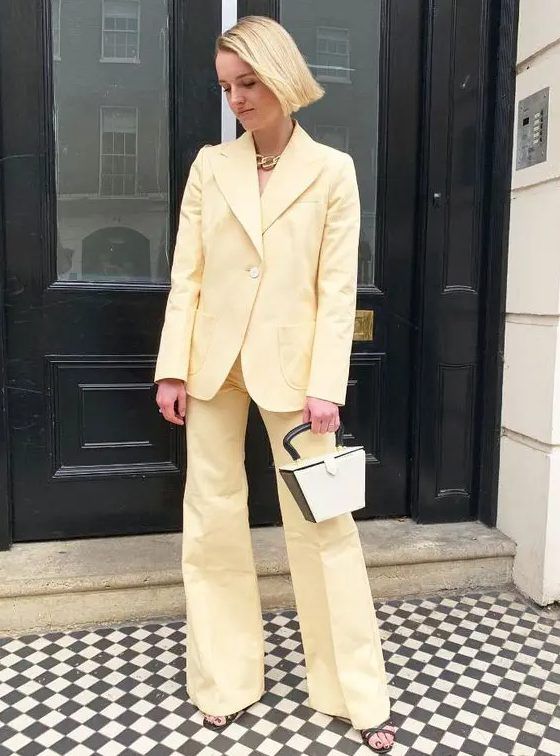 Light yellow pantsuit with flare pants, no top underneath, two-tone bag, strappy shoes, and a chunky chain necklace