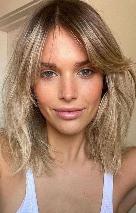Long and messy blonde bob with a darker root and side bangs, with a lot of dimensional texture, a super chic idea