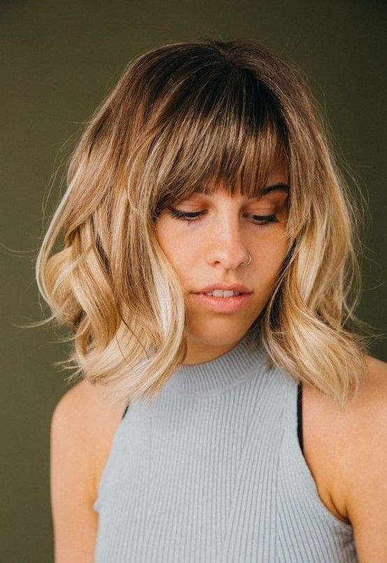 Long blonde bob with a darker root and waves plus wispy bangs for an effortlessly chic and cool look
