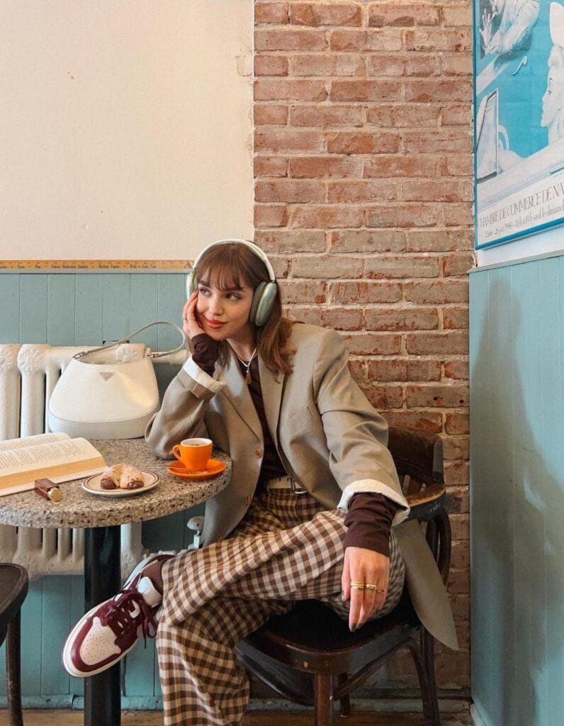 A woman sitting at a cafe table next to a brick wall wearing a casual Dark Academia outfit with a beige oversized blazer, a long-sleeve dark brown top, brown and white plaid trousers, and burgundy sneakers while wearing over-ear headphones.
