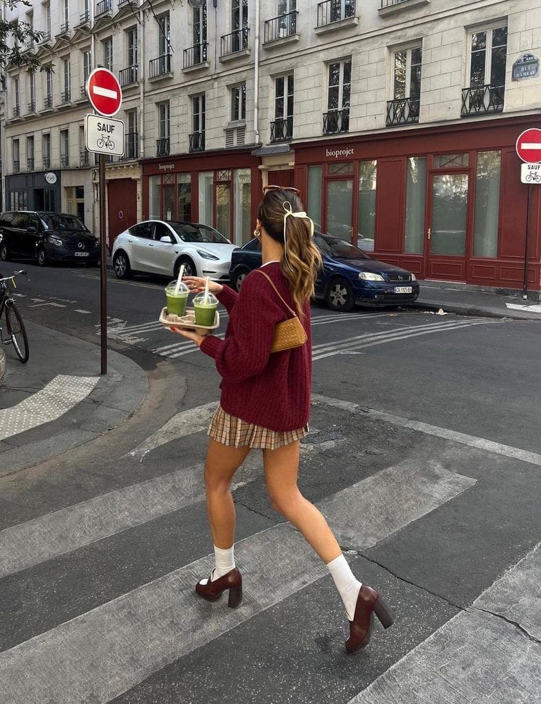 A woman walking across a city street wearing a casual Dark Academia style outfit with a burgundy knit sweater, plaid pleated mini skirt, white socks, and brown block heel loafers.