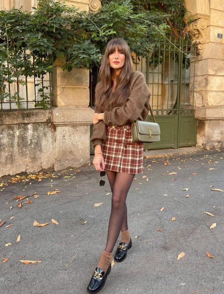 A woman standing outdoors in a Dark Academia outfit featuring a fuzzy brown mohair sweater, a red and brown plaid wool mini skirt, sheer black tights, and black leather chain loafers.