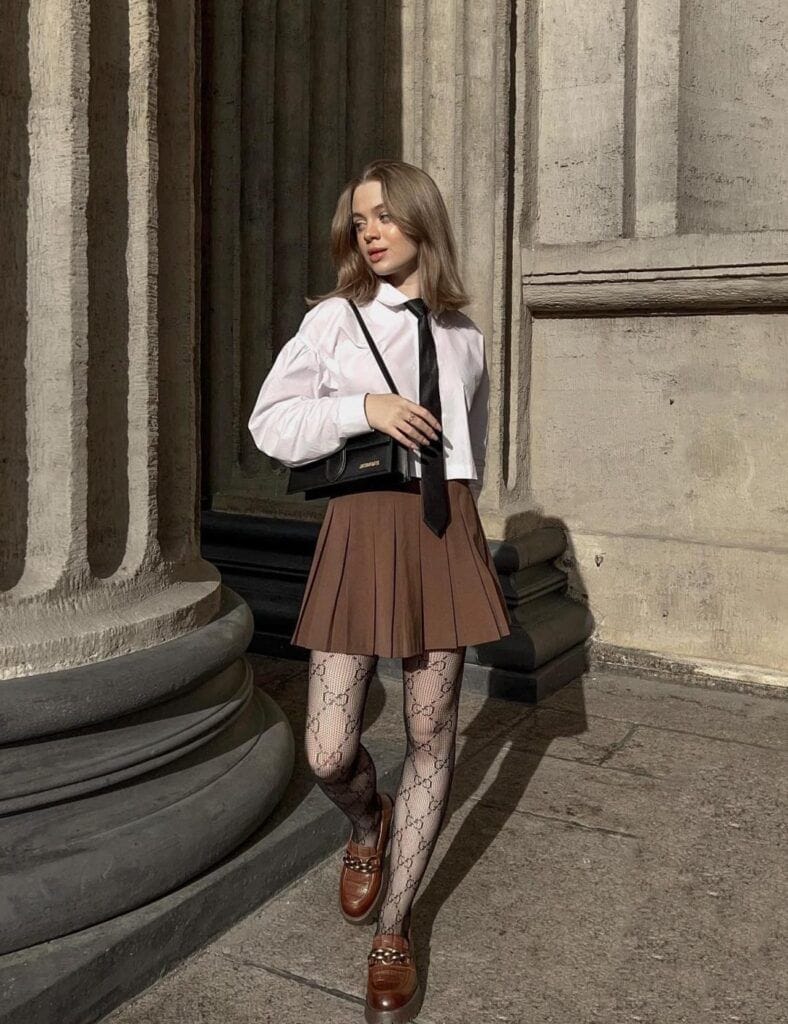 A woman standing near historic stone pillars wearing a classic school-inspired Dark Academia outfit with a crisp white long-sleeve collared shirt, a black necktie, a brown pleated mini skirt, patterned black tights, and brown leather loafers.