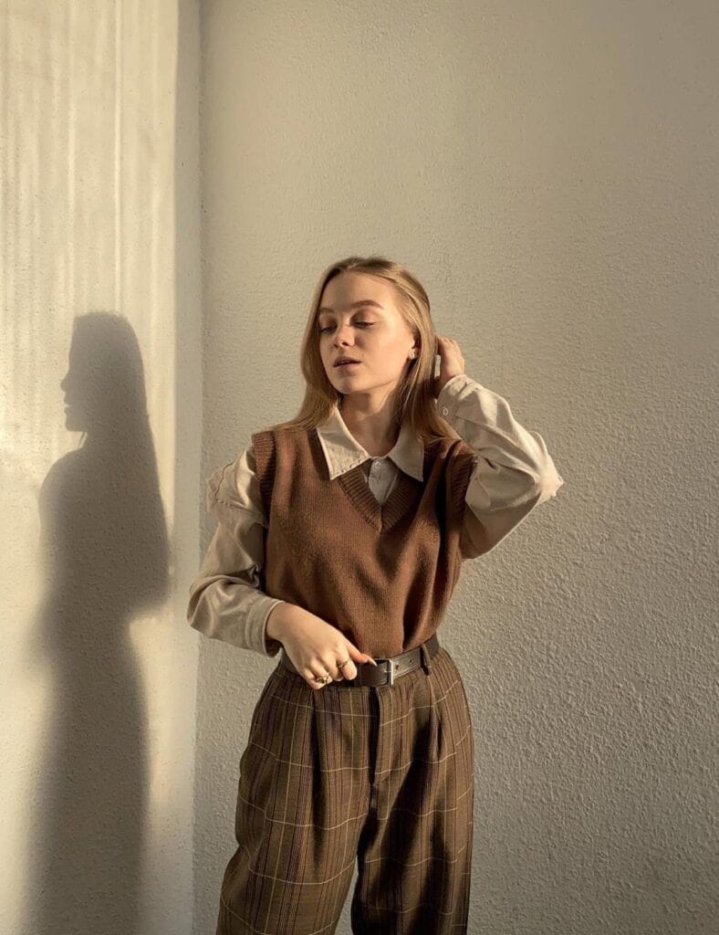 A woman standing in warm sunlight wearing a classic Dark Academia outfit featuring a brown knit V-neck sweater vest layered over a beige collared button-up shirt, paired with high-waisted plaid pleated trousers and a leather belt.