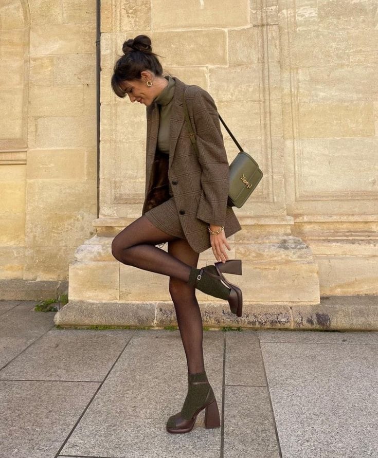 A woman standing by a historic stone building wearing a Dark Academia outfit featuring a brown plaid tweed oversized blazer, matching mini skirt, olive green turtleneck, sheer black tights, and chunky brown platform heels.