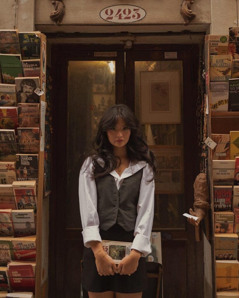 A woman standing at the entrance of a vintage bookstore surrounded by shelves of old books, wearing a Dark Academia outfit with a tailored charcoal grey waistcoat vest layered over an oversized white collared button-up shirt and a black skirt.