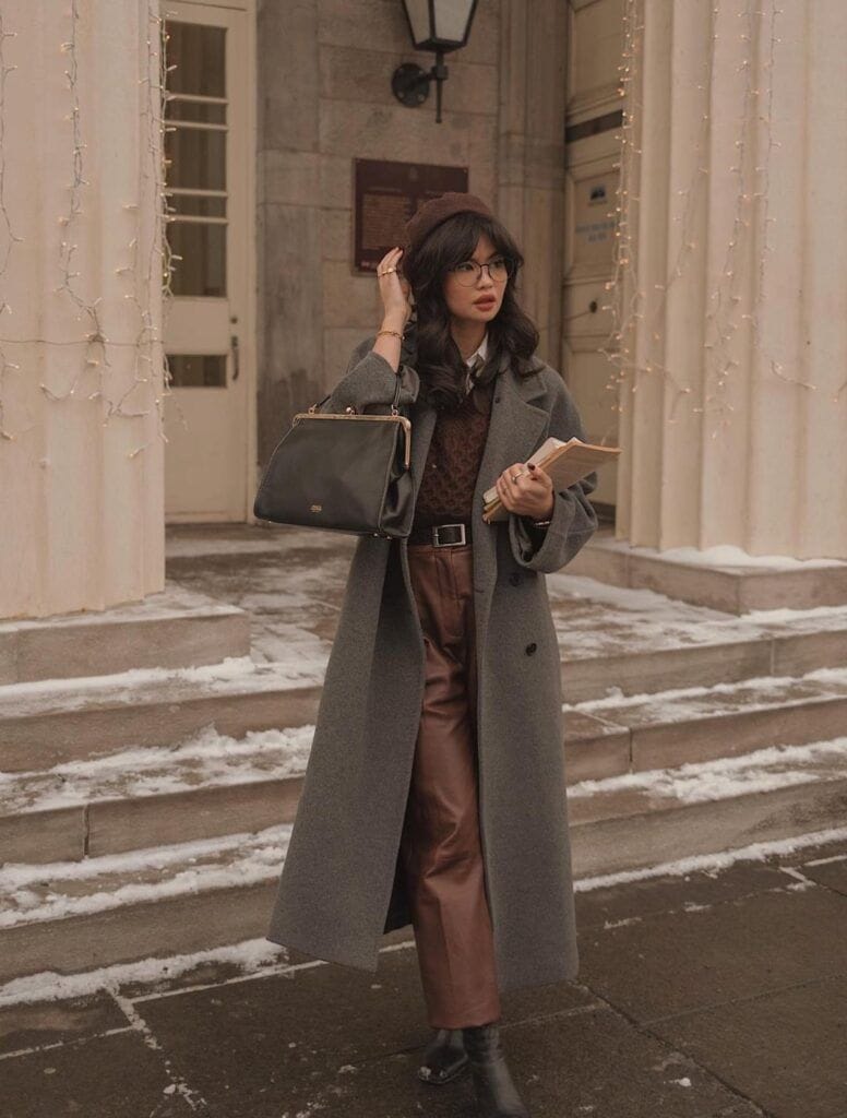 A woman with glasses and a brown beret standing on light snowy steps holding books, wearing a winter Dark Academia outfit with a long grey wool overcoat, a brown cable-knit sweater over a collared shirt, brown wide-leg trousers, and a black leather handbag.