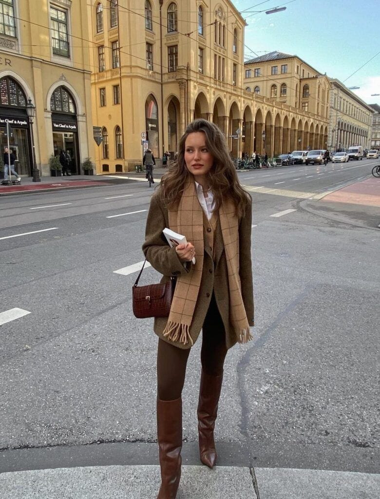A woman standing on a city street holding a book, wearing a layered Dark Academia autumn outfit with a brown wool blazer, a matching vest over a white button-up shirt, brown trousers, knee-high brown leather boots, and a beige windowpane checkered scarf.